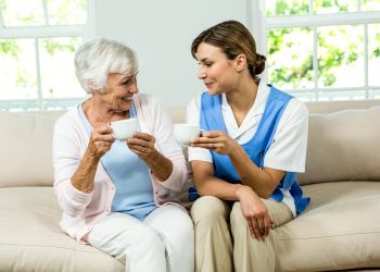 Ältere Frau sitzt mit ihrer Haushaltshilfe auf einem Sofa, beide halten eine Kaffeetasse – Haushaltsunterstützung für Angehörige in Kiel